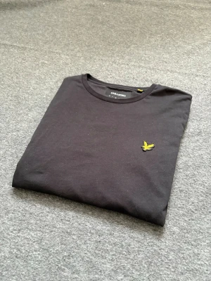 Svart t-shirt från Lyle & Scott - Svart t-shirt från Lyle & Scott med klassisk rund hals och gul broderad logga på bröstet. Tillverkad i mjuk bomull och har en normal passform. Perfekt för en stilren och enkel look.