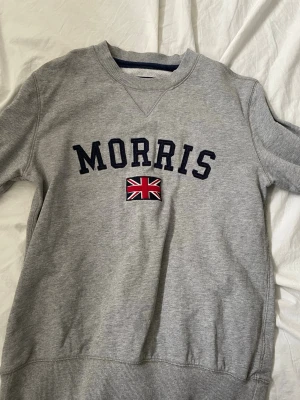 Grå sweatshirt från Morris - Snygg grå sweatshirt från Morris med mörkblå text och en broderad Union Jack-flagga på bröstet. Tröjan har rund halsringning, ribbade muddar och är tillverkad i mjukt bomullsmaterial. Perfekt för en avslappnad och stilren look.
