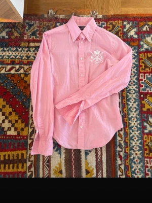 Rosa skjorta från Ralph Lauren - Säljer en snygg rosa skjorta från Ralph Lauren med vit broderad logga på bröstet. Skjortan har klassisk krage, knappar framtill och långa ärmar. Perfekt för dig som vill ha en stilren och trendig look.