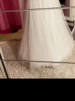 Vit axelbandslös långklänning - Säljer en elegant vit långklänning med axelbandslös design och vacker spetsdetalj vid bysten. Klänningen har ett svepande fall i flera lager tyg som ger en romantisk känsla. Perfekt för speciella tillfällen när du vill känna dig extra fin.Längd på hela klänningen ca 140 cm  Mått armhålan till armhålan 48 cm  2 underkjolar ingår.  Brudslöjan är ca 150 cm lång och har pärlor.