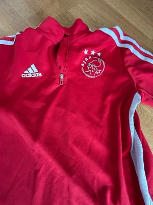 Röd Ajax träningströja från Adidas - Röd träningströja från Adidas med Ajax Amsterdam-logga och tre stjärnor på bröstet. Tröjan har halv dragkedja, vita detaljer och klassiska Adidas-ränder på axlarna. Perfekt för fotbollsträning eller sportiga dagar.