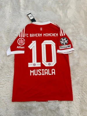 Bayern München Musiala matchtröja - Röd och vit Bayern München matchtröja från Adidas med Musiala och nummer 10 på ryggen. Tröjan har Champions League-märke på ärmen, klubbmärke och sponsorloggor framtill samt klassiska Adidas-ränder på axlarna. Perfekt för fotbollsfans!