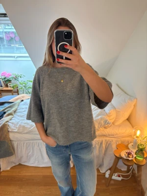 Grå stickad tröja med halvlånga ärmar - Mysig grå stickad tröja med avslappnad passform och halvlånga ärmar. Tröjan har en enkel, rak design och är perfekt att slänga på sig över ett par jeans. Materialet ser ut att vara mjukt och skönt, perfekt för kyligare dagar.