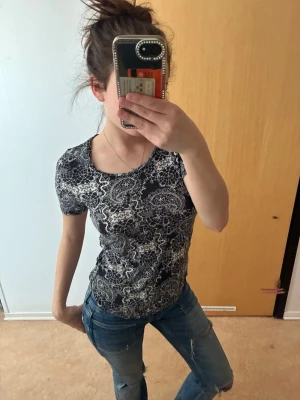 Svart paisleymönstrad t-shirt - Snygg svart t-shirt med vitt paisleymönster och korta ärmar. Figurnära passform och rund halsringning. Perfekt för dig som gillar unika mönster och vill sticka ut lite extra. Materialet känns mjukt och stretchigt, vilket gör den bekväm att bära hela dagen.