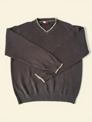 Brun stickad tröja Tommy Hilfiger - Mörkbrun stickad tröja från Tommy Hilfiger med beige ribbade detaljer vid hals, ärmslut och nederkant. V-ringad modell med diskret logga på bröstet. Perfekt för lager på lager och klassisk stil. Har ett pyttelitet hål i nacken men märks inte.