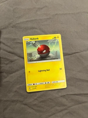 Pokémonkort Voltorb - Samlarobjekt! Ett Pokémonkort med Voltorb-motiv, gul ram och illustration av Voltorb med blixtar i bakgrunden. Kortet har attacken Lightning Ball och HP 50. Perfekt för dig som gillar Pokémon eller samlar på kort.