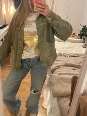 Grön jeansjacka från Zara - Säljer en olivgrön jeansjacka med avslappnad passform och stora utanpåfickor framtill. Jackan har klassisk krage, knappstängning och synliga sömmar för en cool vintagekänsla. Perfekt att slänga över en t-shirt för en chill look.