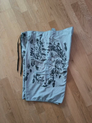 Gråa Billabong shorts med tryck - Snygga gråa shorts från Billabong med svart och vitt grafiskt tryck på båda sidor. Klassisk modell med snörning i midjan och ficka med lock på sidan. 