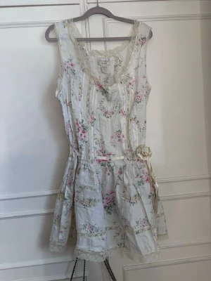 Blommig vit kortklänning med spets - Söt vit kortklänning med blommigt mönster i rosa och grönt. Klänningen är ärmlös, har spetsdetaljer vid kanterna och ett band i midjan som kan knytas till rosett. Perfekt för sommarens varma dagar. Hör av er vid frågor💕