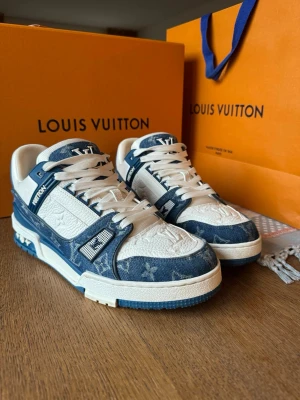 Louis Vuitton blåvita sneakers - Exklusiva Louis Vuitton sneakers i blått och vitt med ikoniskt monogrammönster. Skorna har denimdetaljer, vita läderpaneler och tydliga LV-loggor på både plös och häl. Snygg design med robust sula och coola kontraster. Perfekta för dig som gillar lyxiga streetwear-vibbar.