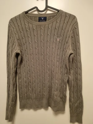 Grön kabelstickad tröja från GANT S - Kabelstickad tröja i beige från GANT med rund halsringning och diskret logga på bröstet. Tillverkad i stretchig bomull för extra komfort. Perfekt för lager-på-lager och snygg till jeans eller kjol. Storlek S