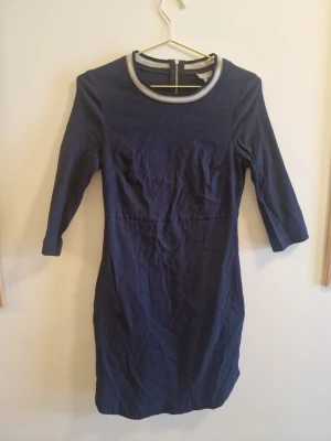 Mörkblå klänning från Oliver Bonas - Mörkblå klänning från Oliver Bonas med trekvartsärm och rund halsringning med silverfärgad detalj. Klänningen har en figurnära passform och dragkedja i ryggen. Perfekt för dig som gillar stilrena och enkla plagg med en twist.