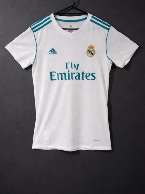 Real Madrid Ronaldo 7 matchtröja - Vit Real Madrid matchtröja från Adidas med turkosa detaljer och tryck. Framsidan har klubbmärke, Adidas-logga och Fly Emirates-tryck. Baksidan har RONALDO och nummer 7. Korta ärmar och klassisk passform, perfekt för fotbollsträning eller support.