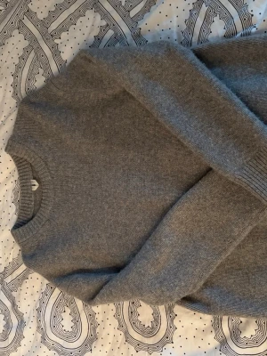 Grå stickad tröja från Arket - Mysig grå stickad tröja från Arket med rund halsringning och ribbade muddar vid ärmslut och hals. Tröjan har en enkel och stilren design som passar perfekt till kalla dagar. Materialet känns mjukt och behagligt mot huden.