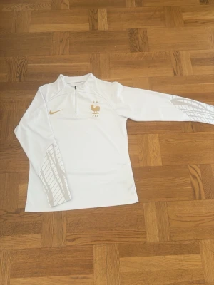 Vit Nike träningströja Frankrike - Snygg vit långärmad träningströja från Nike med Frankrikes landslagsmärke i guld på bröstet och guldfärgad Nike-logga. Tröjan har halv dragkedja och diskreta rutmönster i grått på ärmarna. Perfekt för fotbollsträning eller sport.