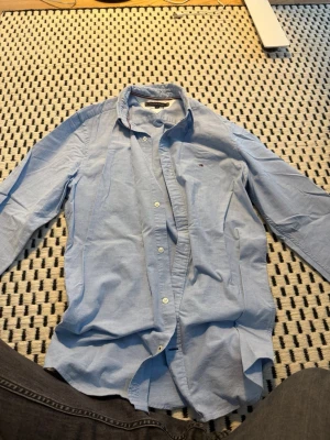 Ljusblå skjorta från Tommy Hilfiger - Klassisk ljusblå skjorta från Tommy Hilfiger med diskret logga på bröstet. Skjortan har långa ärmar, knappar framtill och är tillverkad i bomull med stretch för extra komfort. Perfekt till jeans eller chinos.