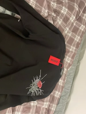 Svart hoodie med ros från HUGO - Svart hoodie från HUGO med stor tryckt röd ros och taggtrådsmotiv på ryggen. Märkets logga syns tydligt och materialet känns mjukt och bekvämt. Perfekt för dig som gillar streetwear med en unik twist.