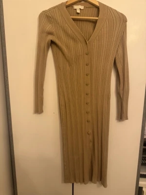 Beige ribbad långklänning H&M - Snygg beige långklänning från H&M med ribbad struktur och v-ringning. Klänningen har långa ärmar och knappar hela vägen framtill, vilket ger en stilren och trendig look. Perfekt för dig som gillar en enkel men elegant stil.