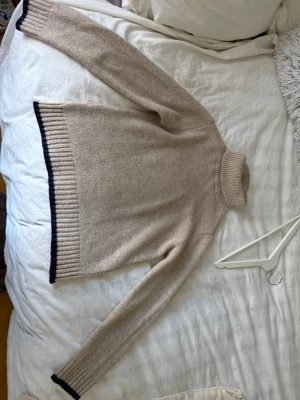 Beige polotröja med svarta detaljer - Stickad beige polotröja med långa ärmar och blåa ribbade kanter vid ärmslut och nederkant. Tröjan har en hög polokrage och är i mjukt material, perfekt för kyliga dagar.