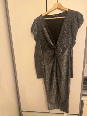 Glittrig silverklänning från Boohoo - Snygg glittrig silverklänning från Boohoo med djup v-ringning, markerad midja och puffiga axlar. Klänningen har lång ärm och en slits framtill. Materialet är stretchigt och skimrande, perfekt för att sticka ut.