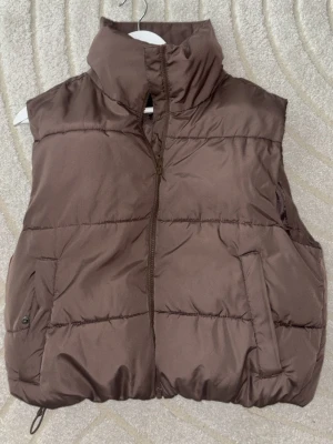 Brun dunväst från Bik Bok - Snygg brun dunväst från Bik Bok i storlek S. Västen har hög krage, dragkedja framtill och två fickor. 