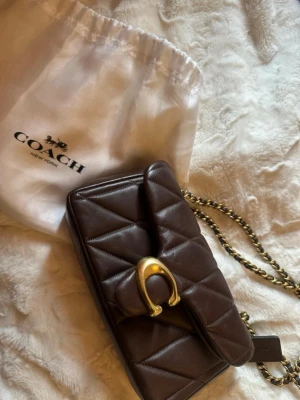 Brun quiltad handväska från Coach - Snygg brun quiltad handväska från Coach med guldfärgad kedjerem och dekorativt spänne framtill. Väskan är tillverkad i mjukt läder och har ett klassiskt lock med magnetlås. Perfekt accessoar för att lyfta din outfit.