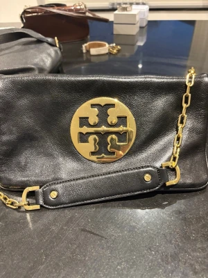 Svart axelväska från Tory Burch - Stilren svart axelväska i läder från Tory Burch med stort guldfärgat logomärke framtill. Väskan har en kedjerem i guld och detaljer i samma färg. Perfekt för dig som gillar klassiska och trendiga accessoarer.