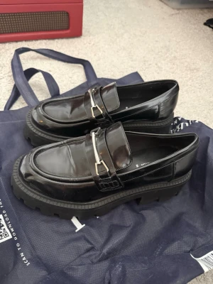 Svarta chunky loafers Pull&Bear - Stilrena svarta loafers från Pull&Bear med chunky sula och dekorativt spänne i metall. Skorna är i glansigt syntetmaterial och har en modern, edgy look som passar perfekt till jeans eller kostymbyxor.