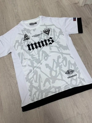 Vit och svart t-shirt från Squadshit - Snygg vit t-shirt från Squadshit med svart och grått grafiskt mönster framtill. Kortärmad modell med svarta detaljer vid ärmslut och nederkant. Tryck och loggor på bröstet och magen ger en cool streetwear-känsla.