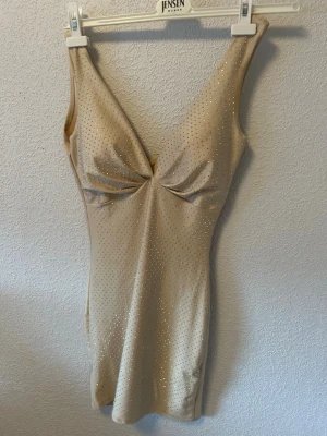 Beige klänning med glitter diamebt - Snygg beige body med djup urringning och twistad detalj vid bysten. Tunt, stretchigt material med små glittrande strass över hela plagget. Perfekt för dig som vill ha något extra under kläderna. Axelbandslös design för en clean look.