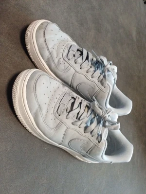 Nike Air Force 1 vita gråa sneakers Storlek 38,5  - Klassiska Nike Air Force 1 sneakers i helvitt läder med perforerade detaljer på tån och vit sula. Skorna har lågt skaft, matchande vita skosnören och den ikoniska swoosh-loggan på sidan. Perfekta för en clean och stilren look. Storlek 38,5 