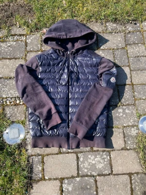 Moncler jacka med quiltad front - Snygg hoodie från Moncler i mörkblått med quiltad, glansig front och mjuka ärmar. Huva med dragsko och dragkedja framtill. Moncler-logga på ärmen och praktisk ficka med knapp. Perfekt för en trendig streetstyle-look.