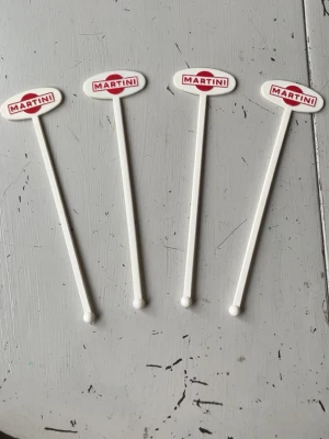 Martini drinkpinnar i plast - Fyra vita drinkpinnar från Martini med logotyp i rött och vitt på toppen. Pinnarna är av plast och har en rundad ände för att röra om drinkar. Perfekt som baraccessoar eller samlarobjekt.