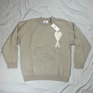 Beige stickad tröja från Ami Paris - Snygg beige stickad tröja från Ami Paris med deras ikoniska vita hjärta och A-logga på bröstet. Tröjan har rund halsringning, ribbade muddar och är tillverkad i mjukt material som känns skönt mot huden. Den är i storlek M och passar ganska baggy. Skriv vid minsta frågor eller funderingar!