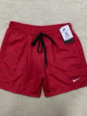 Röda Nike shorts med snörning - Snygga röda shorts från Nike med svart snörning i midjan och vit logga på benet. Perfekta för sommarens aktiviteter och har en sportig look. Tillverkade i ett lätt och bekvämt material.