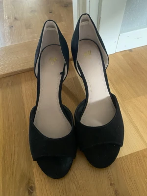 Svarta pumps från H&M med öppen tå - Säljer ett par stilrena svarta pumps från H&M i storlek 39. Skorna har öppen tå och klack, samt en elegant skärning på sidan. Materialet är mockaimitation som ger en mjuk känsla och snygg finish. Perfekta för dig som vill ha en klassisk och trendig look.