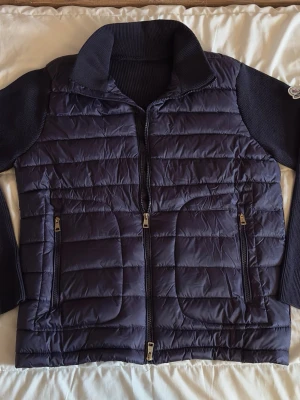 Moncler Cardigan - Snygg mörkblå pufferjacka från Moncler med stickade ärmar och krage. Jackan har två dragkedjeförsedda fickor framtill och quiltad front i glansigt material. Moncler-logga på ärmen. Perfekt för kalla vinterdagar.