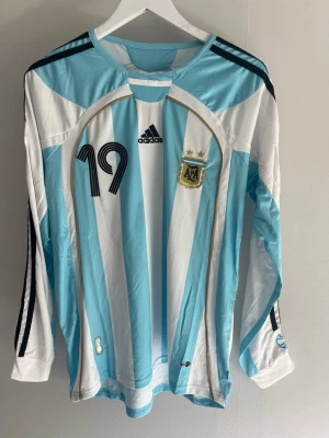 Argentina Messi långärmad matchtröja - Långärmad Argentina fotbollströja med ljusblå och vita ränder, nummer 19 och Messi-tryck på ryggen. Tröjan har svarta ränder på ärmarna och officiellt VM-märke på ärmen. Perfekt för fotbollsfans!