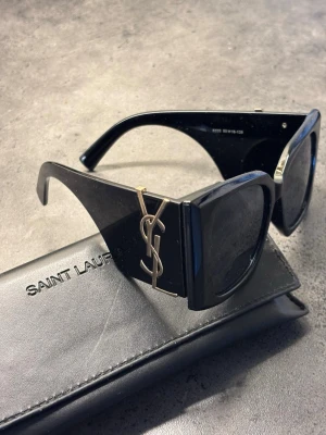 Svarta solglasögon - Snygga svarta solglasögon med breda bågar och ikoniskt emblem i guld på sidorna. Glasögonen har en markerad, fyrkantig form och kommer med originalfodral. Perfekt accessoar för att lyfta din stil.