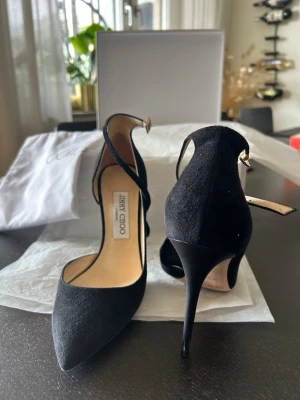 Jimmy Choo Svarta pumps  - Snygga svarta pumps från Jimmy Choo i mocka med spetsig tå och smal hög klack. Skorna har en elegant rem med spänne runt vristen för extra stilpoäng. Perfekta för dig som vill ha en klassisk och trendig look.