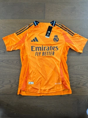 Real Madrid orange matchtröja Mbappé - Snygg orange Real Madrid matchtröja från Adidas med svarta detaljer och tryck. Framsidan har klubbmärke och sponsorlogga, baksidan har Mbappé och nummer 9. Tillverkad i lätt och ventilerande material, perfekt för fotbollsträning eller match.