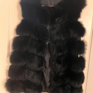 Svart fluffig pälsväst - Säljer en svart pälsväst med riktigt fluffig och mjuk känsla. Västen har en rak form och är tillverkad i pälsmaterial som ger en lyxig vibe. Perfekt att slänga över en hoodie eller tröja för att höja din outfit. Färgen är djup svart och designen är klassisk utan synliga detaljer.