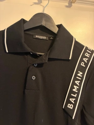 Svart pikétröja från Balmain Paris - Snygg svart pikétröja från Balmain Paris med vita detaljer och logga på axeln. Tröjan har klassisk krage, knappar framtill och korta ärmar med vita ränder. Perfekt för en trendig och sportig look.