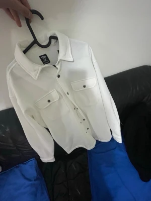 Vit overshirt från FSHN - Stilren vit overshirt från FSHN med klassisk krage och två stora bröstfickor med knappar. Rak passform och svarta knappar framtill ger en clean look. Perfekt att slänga över en hoodie eller t-shirt för en chill vibe.