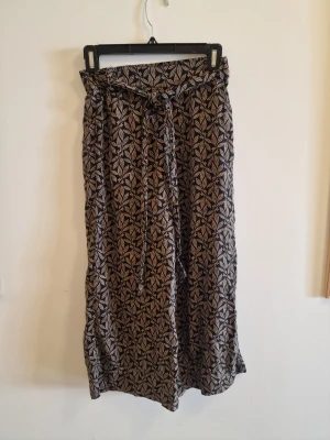 Svarta byxor med bladprint - Snygga svarta tygbyxor med beige bladmönster och knytskärp i midjan. Vida ben och avslappnad passform, perfekta för en chill och trendig look. Materialet känns lätt och luftigt, perfekt för varma dagar.