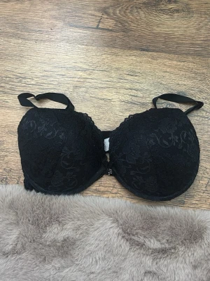 Svart spets-bh med vaddering - Snygg svart bh med vadderade kupor och spetsdetaljer. Axelbanden är justerbara och bh:n har en liten rosett framtill. Perfekt för dig som gillar en klassisk och elegant look. Bella push up storlek E80. Nypris 249kr