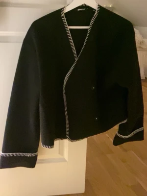 Svart kofta med vita detaljer - Svart kofta med vit dekorationssöm längs kanterna och ärmsluten. Koftan har en rak, boxig passform och knäpps med dolda knappar framtill. Perfekt för lager-på-lager och ger en stilren look. En av knapparna funkar inte men det är inget som märks🤗