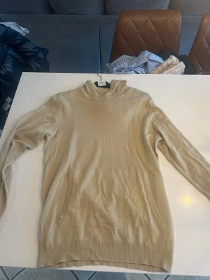 Beige tröja från J.Lindeberg - Stilren beige tröja från J.Lindeberg med lång ärm och ribbade muddar. Tröjan har en högre krage och är tillverkad i ett mjukt, tunt material som passar perfekt för lager-på-lager. Enkel och snygg design som funkar till många outfits.