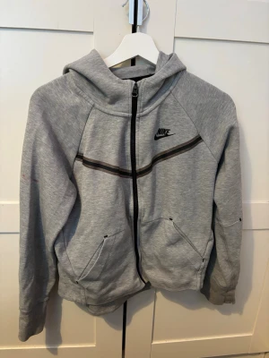 nike tech fleece - Snygg grå hoodie från Nike i storlek XL med svart logga på bröstet och hel dragkedja framtill. Hoodien har två fickor, huva och en svart rand över bröstet. Perfekt för en sportig och avslappnad stil.