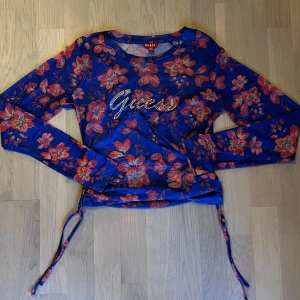 Blommig långärmad topp från Guess - Snygg långärmad topp från Guess i blått med blommigt mönster i rött och rosa. Toppen har glittrande Guess-logga framtill och snörning i sidorna för en cool detalj. Perfekt för dig som vill sticka ut med färg och mönster.